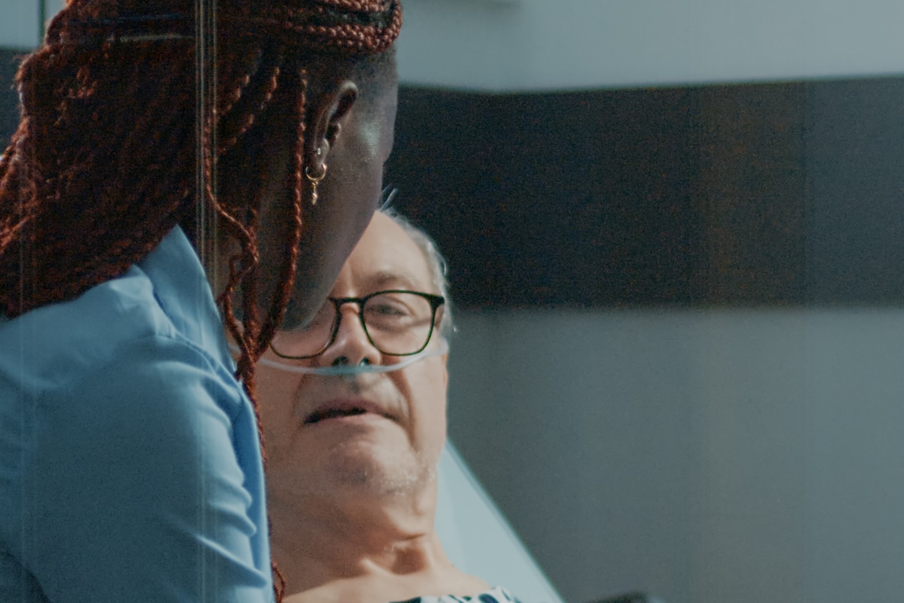 A caregiver sits at the bedside of an older man in hospital, offering comfort